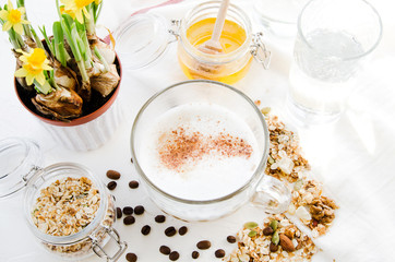 Top view healthy breakfast with cappuccino coffee, cinnamon, granola, muesli, cereal, honey, drizzlier, glass of water, daffodil flowers on a white background. Food, spring concept.