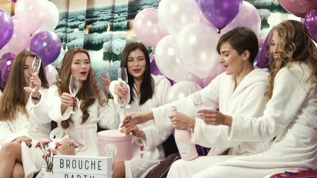Beautiful girls in white robes opening champagne bottle drinking champagne at the opening party. Women celebrating at the beauty salon. Champagne glasses