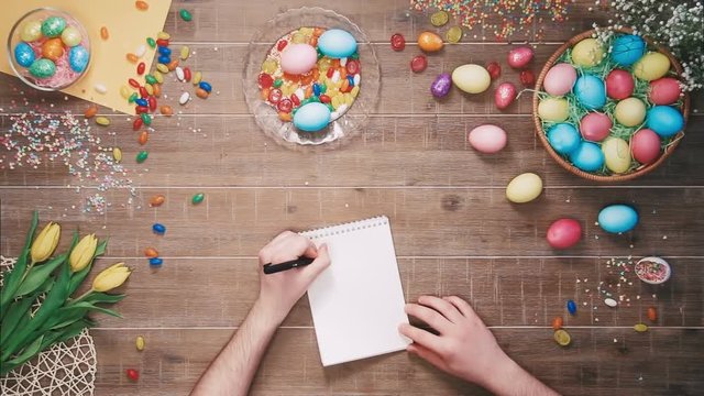 Man writing easter recipe in the notebook on table decorated with easter eggs. Top view