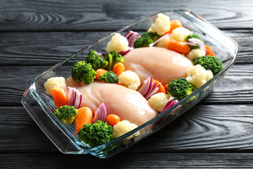 Raw chicken breasts with vegetables in glass baking dish on wooden table