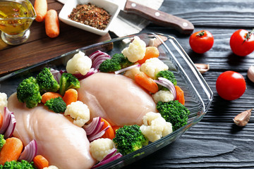 Raw chicken breasts with vegetables in glass baking dish on wooden table