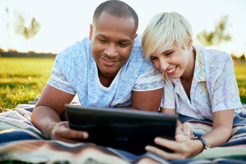 Mixed race couple of millennial in a grass field looking at a digital tablet and reading for their...