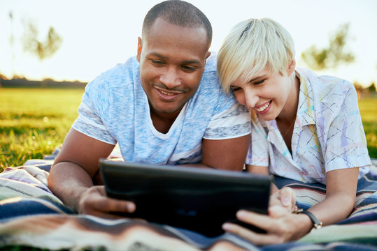 Mixed Race Couple Of Millennial In A Grass Field Looking At A Digital Tablet And Reading For Their Next School Paper