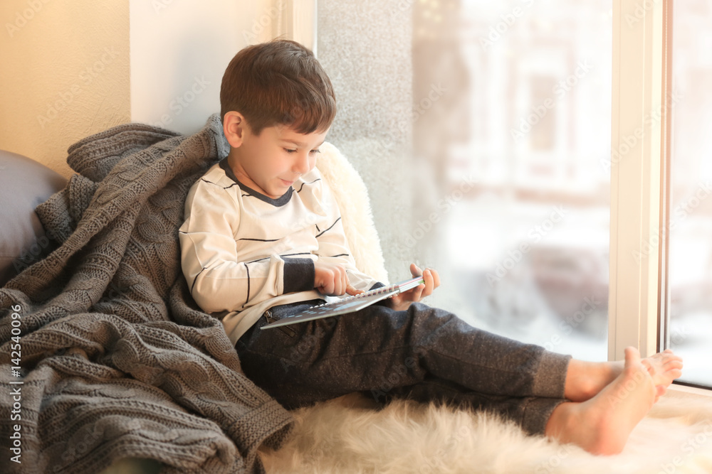 Wall mural Cute little boy reading book while sitting on window sill at home