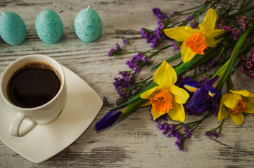 Candles in the form of Easter eggs on a white wooden background. Cup of strong aromatic coffee and beautiful bouquet of spring flowers.