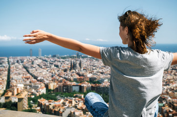 Young happy woman sits back to the camera with raised hands. Traveler enjoying the cityscape of mountain, Barcelona, Catalonia, Spain - obrazy, fototapety, plakaty