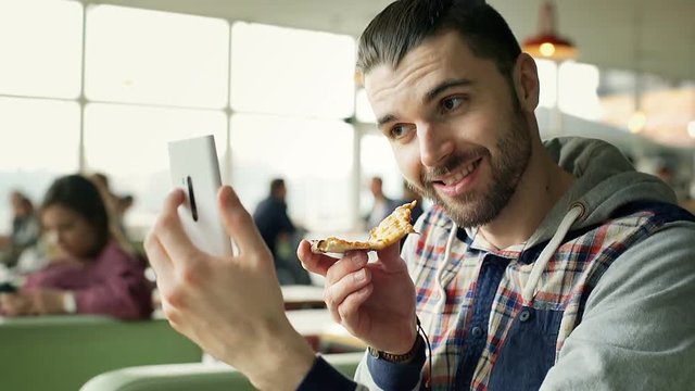 Handsome man doing selfies on smartphone and holding slice of pizza, steadycam shot
