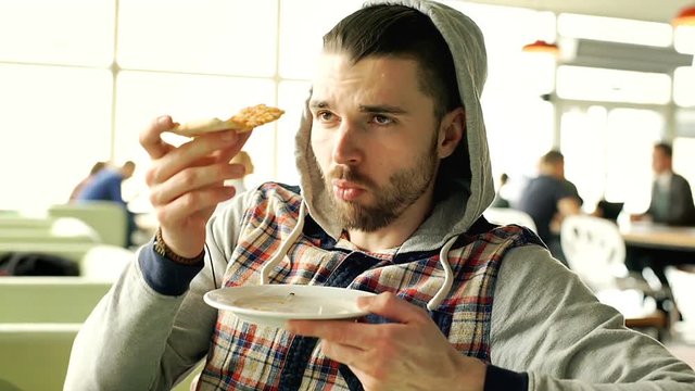 Handsome man wearing hood and eating pizza while chatting with someone, steadycam shot
