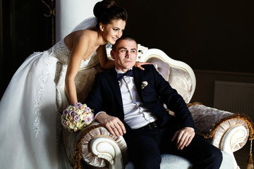 Bride bends over groom while he sits on large armchair