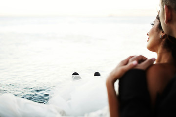 Groom holds his hand on bride's shoulder while they rest on ocean shore