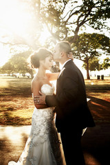 Summer sun shines over groom kissing bride's head before green trees
