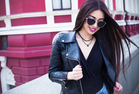 Outdoor lifestyle fashion portrait of young stylish hipster woman walking on street,wearing cute trendy outfit.Young woman with long dark hair  smiling to camera in city on city bulding background.