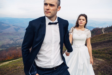 Confident groom holds bride's hand leading her on windy hill