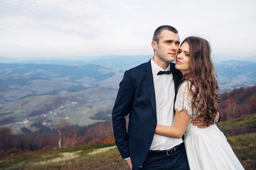 Tall groom in black suit holds bride tender standing under deep autumn sky