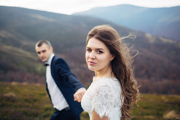 Wind blows bride's brown hair while she climbs hill together with groom
