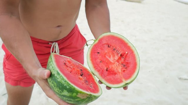 Young Handsome Vegan Man Breaks Watermelon into Two Halves by Strong Hands on the Beach. Thailand. HD Slowmotion.