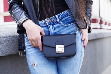 Close up fashion mag of woman sitting outdoor details, vintage black biker leather jacket , ,small bag,purse, trendy blue jeans.Stylishgirl posing on the street, trendy outfit.