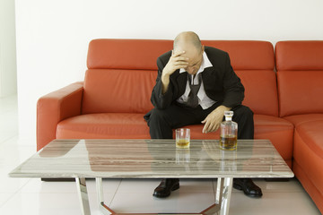 Desperate and depressed man with black suit spending time with bottle of whiskey, result of prolonged stress and frustration, symbol of depression, burnout and loneliness, red sofa background
