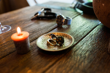 Bronze plate with men’s rings on a wooden table with burning candles. Men’s accessorizes, necktie, watch