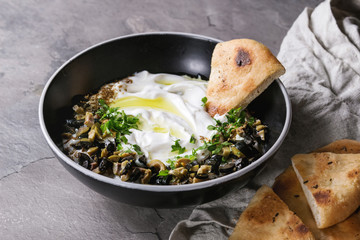 labneh middle eastern lebanese cream cheese dip with olive oil, salt, herbs, olives tapenade served in black bowl with traditional pita bread over gray texture metal background.