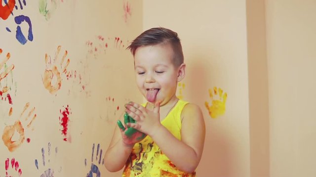 Little boy in yellow sleeveless shirt showing tongue. His hands are dirty in colors. He is living his handprints on the wall. Slowmotion