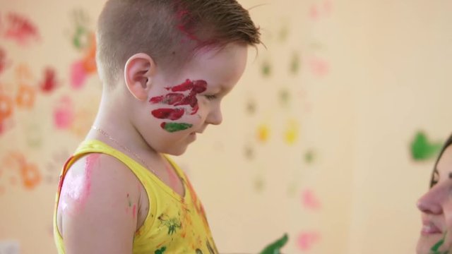 Slow motion and close-up shot of a happy little boy giving tight hugs to his beloved mother while they are playing and leaving their colorful fingerprints on the wall