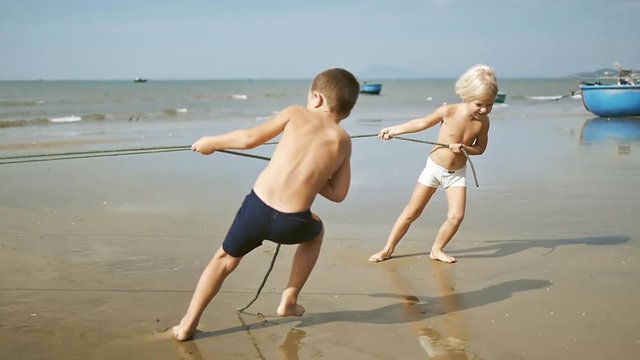 Cute adorable happy children pulling a rope on the tropical sandy beach on sea background.  Having fun on summer vacation.  Helping fisherman. Slow motion