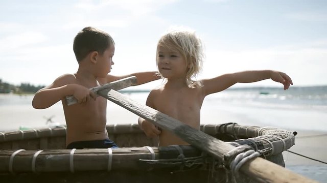 Cute adorable happy children having fun on the sand beach on blue sky and sea background in summer  vacation. Rowing oar sitting in the fishing boat. Playing fishermen, sailors. Vietnam. Slow motion