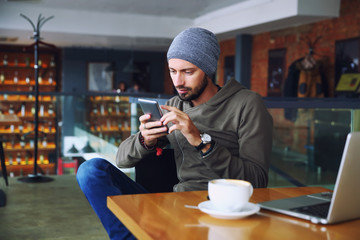 Young handsome hipster man with beard sitting in cafe talking mobile phone, holding cup of coffee and smiling. Laptop on wooden table. - Powered by Adobe