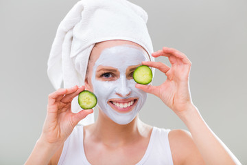Young woman with facial mask holding slices of cucumber on gray background.