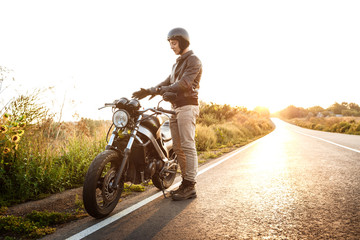Fototapeta premium Young handsome man posing near his motorbike at countryside road. Flare sunlight background.