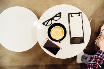 Top view of glasses, cup of cappuccino, clipboard with blank paper and smartphone  in modern cafe...