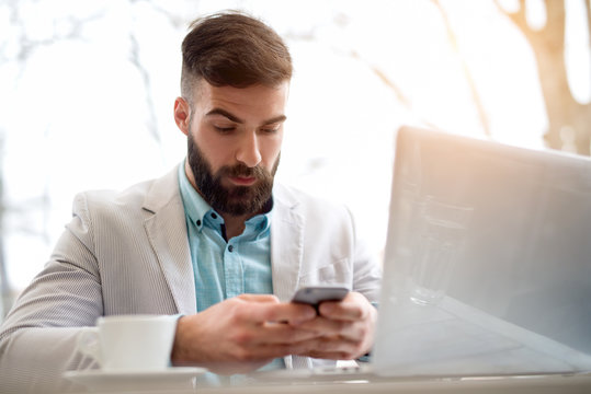 Handsome bearded man in stylish white and blue suit works on computer checking emails on smart phone.
