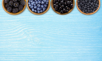 Black and blue berries. Blackberries, blueberries, currants and blueberries in a wooden bowls on a blue background. Berries at border of image with copy space for text. Top view.