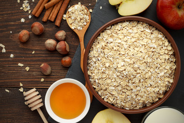 Ingredients for healthy breakfast: rolled oat flakes, milk, honey, hazelnut, cinnamon on dark rustic wooden table.