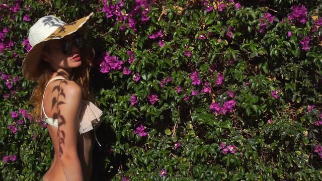 Sexy young blonde lady in straw hat and sunglasses in the blooming garden with magenta flawers summer time