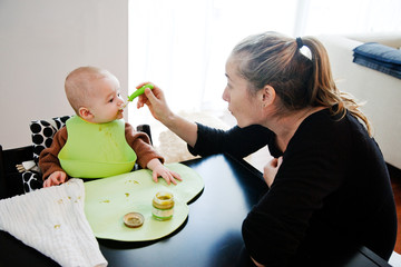 6 month old baby boy sitting in high chair eating with bib being fed by mom