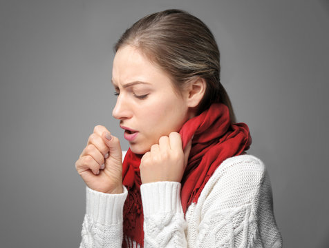 Young woman in warm scarf coughing on gray background