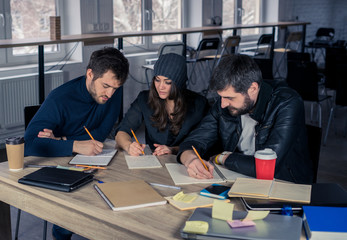 Young students writing in notepads or notebooks in lecture hall. Working environment with laptop, coffee, notepads and stationery.