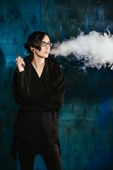 A girl with glasses vaping and releases a cloud of vapor. Model in a black vaper smoke vaporizer on a turquoise background