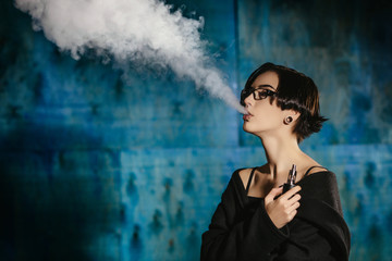 A girl with glasses vaping and releases a cloud of vapor. Model in a black vaper smoke vaporizer on...