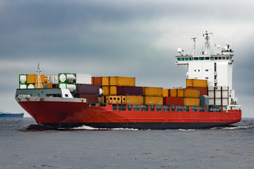 Red cargo container ship sailing from Baltic sea in cloudy day