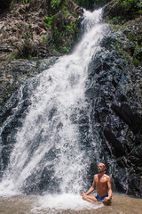 Fototapeta na wymiar Man practicing yoga among waterfall. Young male sitting in lotus pose on rock with waterfall streams near in tropical forest.