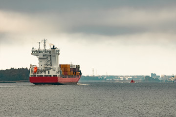 Fototapeta premium Red cargo container ship entering the port of Riga in cloudy day