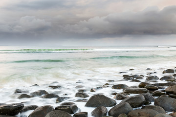 A storm passes on the horizon as waves wash over a boulder strewn beach in this seascape scene.
