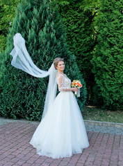 Beautiful light brown bride play with veil on the wind. Attractive girl standing in the park on their wedding day