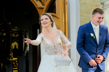 Emotional bride is throwing a candies next to the church. Attractive, smiles bride keeps basket...