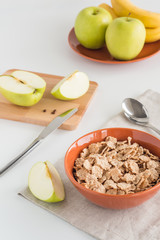 Cornflakes in bowl with fresh apples cut in pieces, bananas, wooden board on white background. Healthy breakfast concept