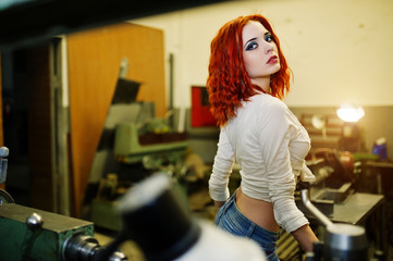 Fototapeta na wymiar Red haired girl wear on short denim shorts and white blouse posed at industrial machine at the factory.