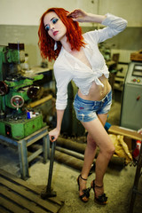 Obraz na płótnie Canvas Red haired girl wear on short denim shorts and white blouse with hammer at hands posed at industrial machine at the factory.